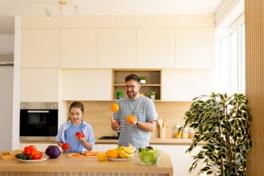 Bir baba ve kızı, parlak bir mutfakta sağlıklı yemekler hazırlarken kahkahalarını paylaşıyorlar. Meyve hokkabazlığı yaparlar, sağlıklı beslenme alışkanlıklarını teşvik eden eğlenceli bir aktiviteye katılırlar..