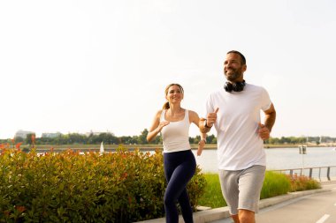 İki birey, canlı bir şehir ortamında, yemyeşil ve köpüklü suyla çevrili nehir kenarındaki yolda ferahlatıcı bir koşunun keyfini çıkarıyor..