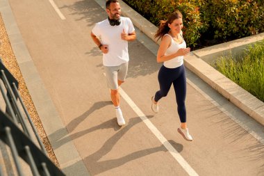 İki koşucu, berrak mavi gökyüzünün altında canlı bitkilerle dolu manzaralı bir yolda ferahlatıcı bir koşunun tadını çıkarıyor..