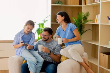 Neşeli bir aile etkileşimi sıcak bir oturma odasında gerçekleşir, bir çocuk mobil bir cihaz kullanırken heyecan gösterir, ebeveynler yakınlarda kahve fincanlarının keyfini çıkarırlar..