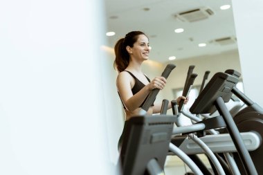 Bir fitness meraklısı, sabah eliptik egzersizi sayesinde modern spor aletleri ve parlak ışıklandırmayla enerji üretiyor..