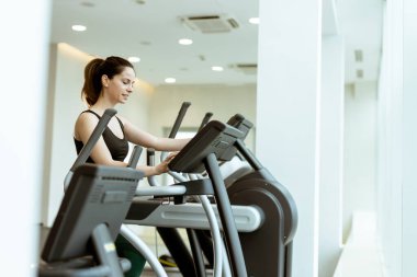 Bir kadın eliptik bir makine üzerinde egzersiz yapıyor, tamamen parlak ve havadar bir spor salonuna odaklanmış, sabahın erken saatlerinde zindelik enerjisini kucaklıyor..