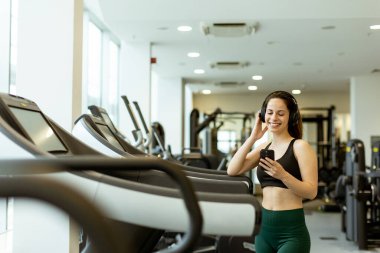 Spor kıyafetleri giymiş bir kadın müzik dinlerken gülümser, spor salonunda spor aletleriyle çevrelenmiş, motivasyonunu arttırır..