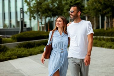 Çağdaş bir şehir parkında, aydınlık ve güneşli bir günde neşe ve arkadaşlığı yansıtan iki kişi huzur içinde yürür..