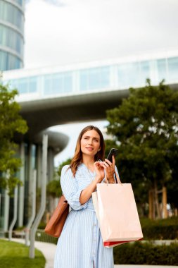 Açık mavi elbiseli bir kadın canlı bir şehir ortamında duruyor, akıllı telefonunu kontrol ediyor ve bulutlu bir gökyüzünün altında alışveriş torbalarını tutuyor..