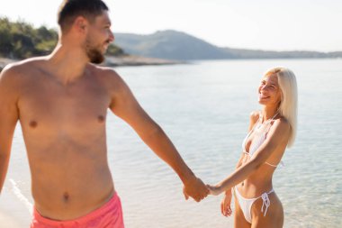 Bir çift, güneşli bir plaj boyunca el ele yürür, yüzlerinde gülümsemeyle. Sıcak güneş suyun üzerinde parıldarken dalgalar yavaşça kıyıya vurur ve mükemmel bir yaz anı yaratır..