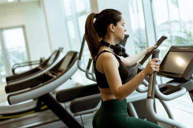 Kararlı bir kadın eliptik bir makine üzerinde enerjik bir egzersiz yapar. Etrafı parlak spor aletleri ve parlak doğal ışık ile çevrilidir..