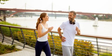 İki koşucu nehir kenarındaki manzaralı bir patika boyunca canlı bir sabah koşusunun tadını çıkarıyor. Neşeli etkileşimleri, yumuşak güneş ışığıyla yıkanan huzurlu ortamın canlı atmosferini güçlendirir..