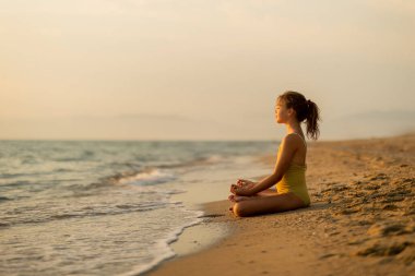 Genç bir kız kumlu sahilde huzur içinde oturur, güneş batarken hafif dalgaların kıyı şeridini kaplamasını seyreder, sakin denizin ve gökyüzünün üzerinde ılık bir parıltı yayar, sükuneti hatırlatır..