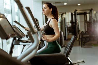 Enerjik bir çalışmayla meşgul olan bir kadın, doğal ışıkla dolu geniş ve modern bir spor salonunda eliptik bir makine üzerinde egzersiz yapıyor..