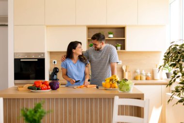 Güneşli ve şık bir mutfakta yemek hazırlarken iki insan neşeli bir anı paylaşıyor. Taze sebzeler tezgahın üzerine seriliyor, aşçılık becerilerini ve beraberliklerini sergiliyorlar..