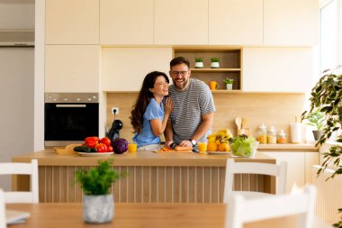 Güneşli bir mutfakta, birkaç kişi yemek hazırlarken kahkahalarını paylaşıyor. Canlı meyve ve sebzelerle çevrili olarak sıcak ve davetkar bir atmosfer yaratırlar..