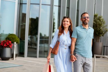 Parlak bir öğleden sonra güneşi, alışveriş torbaları taşıyarak, mağazaları keşfederek geçirdikleri günün heyecanını hissederek, el ele yürürken neşeli bir çifti aydınlatıyor..