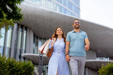 Mutlu bir çift, açık mavi bir gökyüzü altında canlı şehir sokaklarını keşfederken, alışveriş torbaları taşıyarak el ele yürür. Modern mimari onların neşeli deneyimlerini geliştirir..