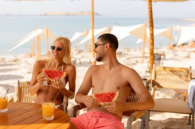 İki arkadaş plaj kenarındaki masada dinleniyor, taze karpuz dilimlerinin tadını çıkarırken aynı zamanda taze içkileri yudumluyorlar. Güneş parlıyor, okyanus kenarında mükemmel bir yaz atmosferi yaratıyor..