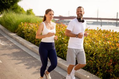 İki arkadaş nehir kenarında spor giysileri içinde enerjik bir koşu yapıyorlar. Güneşin altında gülücükler ve kahkahalar paylaşıyorlar, sağlıklı bir yaşam tarzını ve dostluğu kucaklıyorlar..