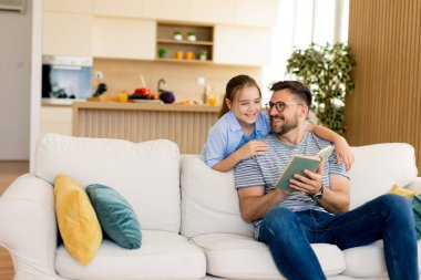 Neşeli bir baba ve kızı, yeşil ve doğal ışıklarla dolu çağdaş bir oturma odasında rahat bir kanepede kitap okurken özel bir bağ kurarlar..