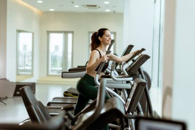 Bir fitness aşığı, şık, aydınlık bir spor salonunda eliptik bir makinede egzersiz yaparken, sağlıklı bir yaşam tarzı ve motivasyonu teşvik ederken ışıl ışıl gülümser..