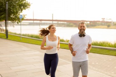Bir kadın ve bir adam nehir kenarında yan yana koşuyor, yüzlerinde kahkahalar var. Açık hava egzersizinin neşesini kucakladıklarında güneş ışığı sudan yansıyor..