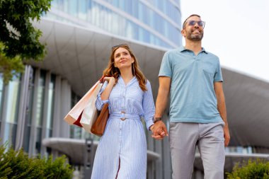 Sıcak bir günde, neşeli bir çift alışveriş torbaları taşıyarak el ele dolaşır ve modern bir binanın önünden geçerken gülümseyerek birlikte şehir hayatının neşesini kucaklarlar..