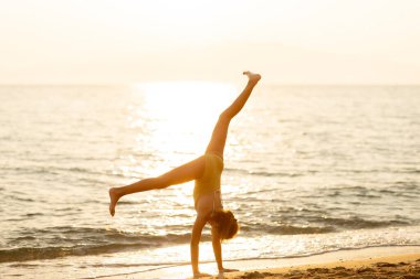 Güneş sakin okyanus dalgalarının üzerinde batarken neşeli genç bir insan kumlu sahilde amuda kalkar ve yaz akşamında bir neşe ve özgürlük anını vurgular..