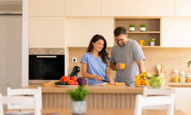 Canlı sebzeler ve taze meyvelerin arasında bir çift, keyifle şık mutfaklarında birlikte yemek hazırlar, keyifli yemek pişirme deneyimi sırasında gülücüklerini ve kahkahalarını paylaşırlar..