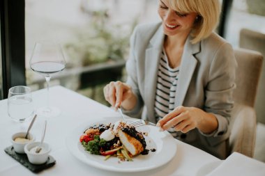 Modern bir restoranda bir masada oturan bir kadın gülümseyerek yemeğinin tadını çıkarıyor. Bir kadeh şarabın tadını çıkarırken güzel, canlı renklerle sunulmuş bir tabağı kesiyor..