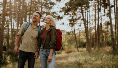 İki yürüyüşçü gülümser ve uzun ağaçlar ve doğanın güzellikleriyle çevrili güneşli bir ormanı keşfederken yukarı bakar..