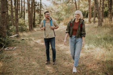 İki arkadaş, uzun ağaçlar ve yeşil yapraklarla çevrili, parlak güneş ışığı altında ağaçlık bir yolda yürüyor. Yürüyüş ve arkadaşlıkla geçen bir günün tadını çıkarıyorlar..