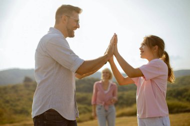 Bir baba ve kız, arka planda manzaranın tadını çıkaran bir kadınla güneşin altında gülümserken el oyunu oynuyorlar. Sahne neşe ve beraberliği yakalar..