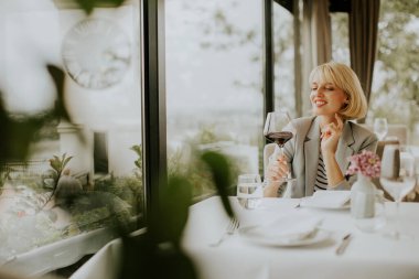Gülümseyen bir kadın bir restoranda şık bir masada elinde bir bardak kırmızı şarapla oturuyor. O şık bir kıyafet giyerken güzel bir açık hava manzarasından hoşlanır..