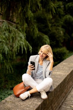 Gündelik giyinmiş genç bir kadın yemyeşil bir ortamda taş bir duvarda oturuyor. Parlak, güneşli bir günde ağaçlarla çevrili akıllı telefonunu kullanırken gülümsüyor..