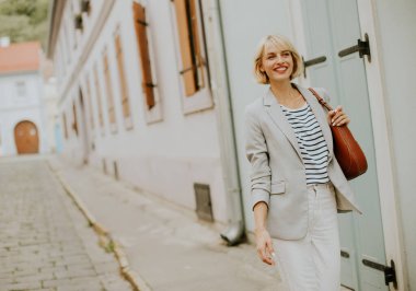 A confident woman walks along a narrow cobblestone street surrounded by beautiful historic buildings. She wears a light blazer and striped shirt, enjoying the sunny weather.