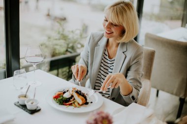 A woman with a bright smile is seated at a table, enjoying a well-presented plate of food in a modern restaurant. She holds a fork and knife, ready to savor her meal.
