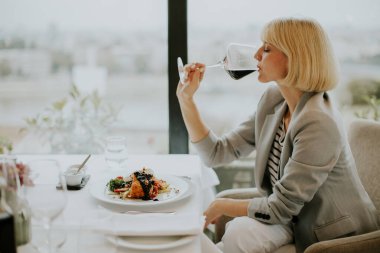 Gri ceketli bir kadın lüks bir restoranda yemek yerken kırmızı şarap yudumluyor. Önünde bir tabak gurme yemeği var ve şehrin ufuk çizgisi pencereden görülüyor..