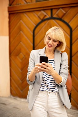 A woman with short blonde hair is smiling while looking at her smartphone. She wears a gray blazer over a striped shirt, standing in front of a stylish wooden wall.