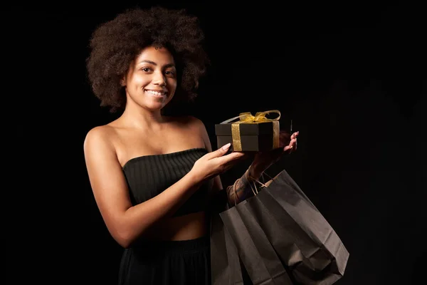 Black Friday sale, gift, present, celebration concept. Smiling afro-american female holding black paper shopping bags and a gift box showing blank credit card, over black background