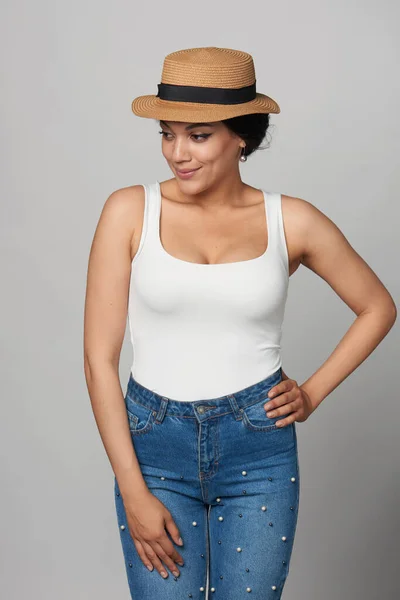Relaxed casual mixed race african american - caucasian woman in jeans and white tank top wearing canotier straw hat, looking down smiling