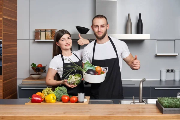 モダンなキッチンで若いカップルの料理サラダ 親指を立ててカメラを見ると — ストック写真