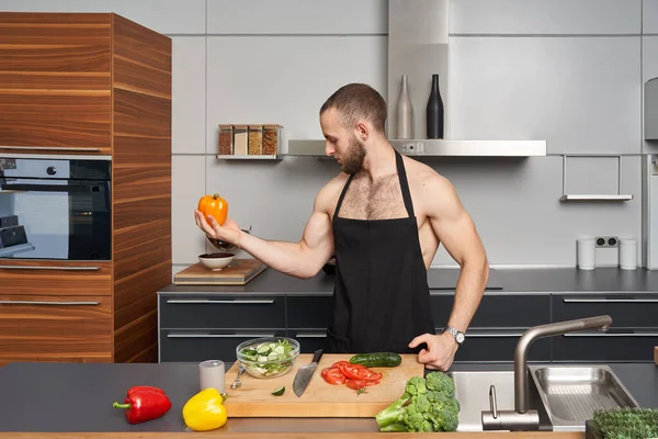 肌肉发达的男人做广告 宣传健康的蔬菜食品 展示他的肌肉 在现代厨房里 穿着大厨围裙的肌肉男在做沙拉 — 图库照片#
