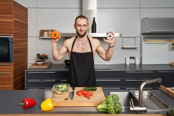 ドーナツと健康的な食べ物の間で選択筋肉のスポーティ男 シェフエプロンを身に着けている愚かな筋肉男料理サラダで現代的なキッチン — ストック写真