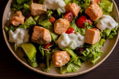 Lezzetli bir Akdeniz salatasına yakın plan. İçinde somon filetosu ve bir tutam tzatziki sosu var. Taze ve lezzetli bir yemek deneyimi sunuyor.