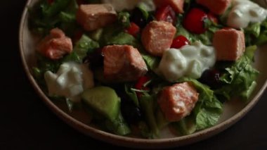 Taze ve sağlıklı bir akşam yemeği deneyimi sunan, somon parçaları ve bir tutam tzatziki soslu lezzetli bir Akdeniz salatasını gösteren yakın çekim bir video.