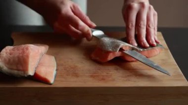 Ahşap bir tahtanın üzerindeki somon filetosunun derisini nazikçe yüzen bir kadının ellerini gösteren ve taze deniz ürünlerinin hazırlıklarını gösteren yakın plan bir video.