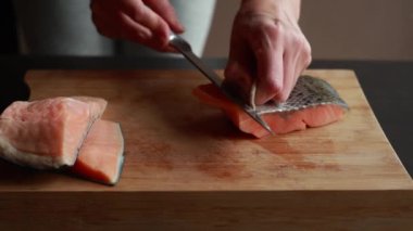 Ahşap bir tahtanın üzerindeki somon filetosunun derisini nazikçe yüzen bir kadının ellerini gösteren ve taze deniz ürünlerinin hazırlıklarını gösteren yakın plan bir video.