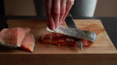 Ahşap bir tahtanın üzerindeki somon filetosunun derisini nazikçe yüzen bir kadının ellerini gösteren ve taze deniz ürünlerinin hazırlıklarını gösteren yakın plan bir video.