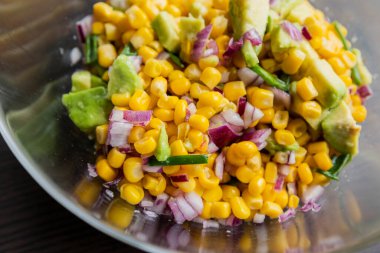  İçinde mısır, kırmızı soğan ve avokado bulunan, limon sosu serpiştirilmiş bir yaz salatasının canlı renkleri, bu yakın planda cam bir kasede sunuluyor.