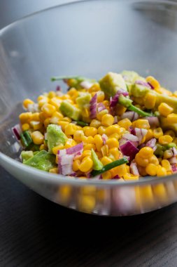  İçinde mısır, kırmızı soğan ve avokado bulunan, limon sosu serpiştirilmiş bir yaz salatasının canlı renkleri, bu yakın planda cam bir kasede sunuluyor.
