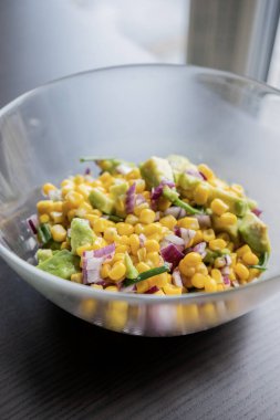 İçinde mısır, kırmızı soğan ve avokado bulunan, limon sosu serpiştirilmiş bir yaz salatasının canlı renkleri, bu yakın planda cam bir kasede sunuluyor.