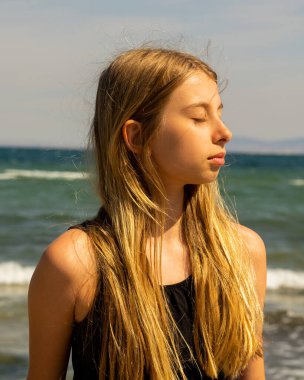 Siyah bir yaz elbisesi giymiş, kumsal arka planında rüzgarda savrulan sarışın bir kızın yakın plan portresi.